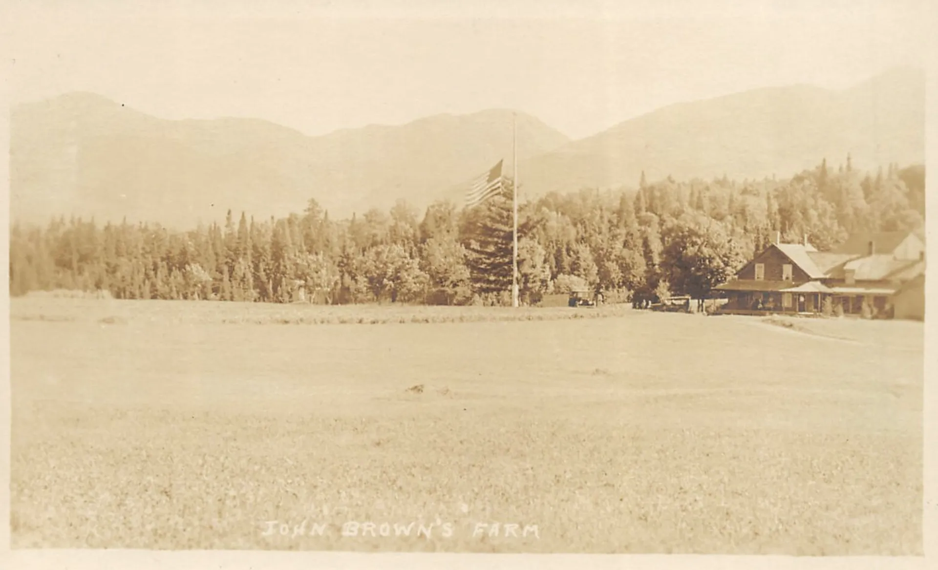 An antique, sepia-toned postcard showing a farmhouse and American flag in front of a mountain range.